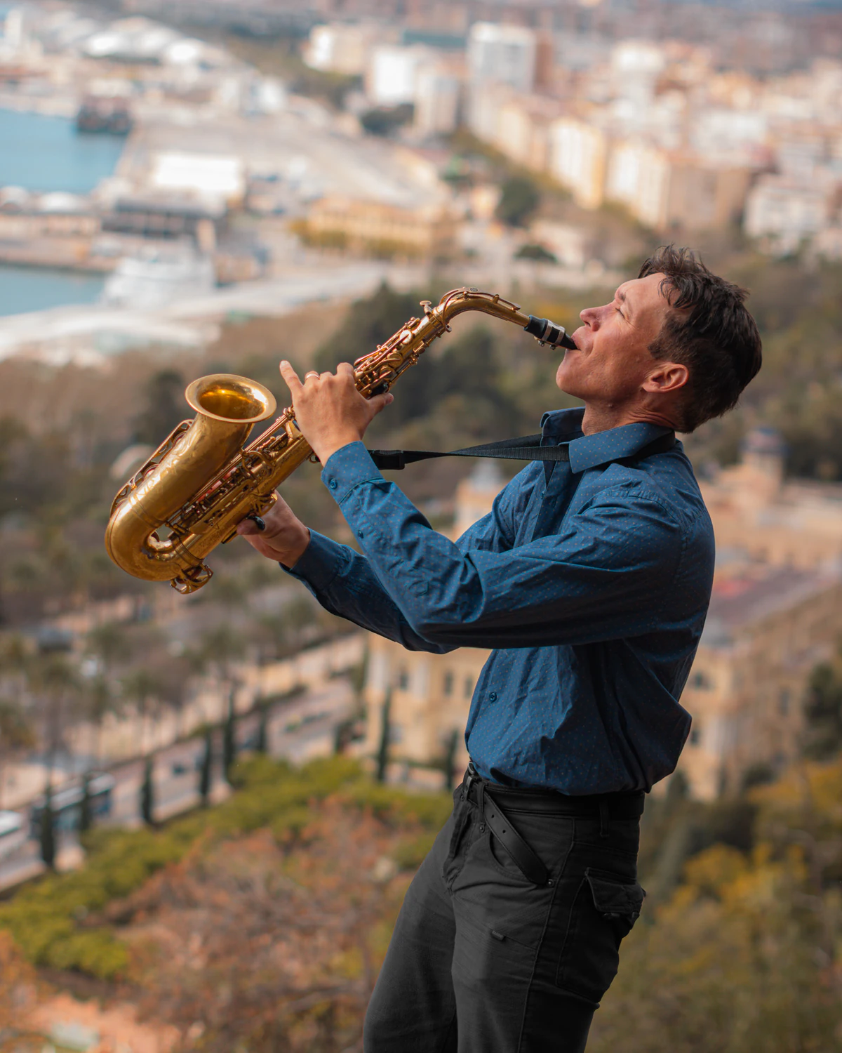 Zirano playing the saxophone at Gibralfaro, with Malaga and the port behind him