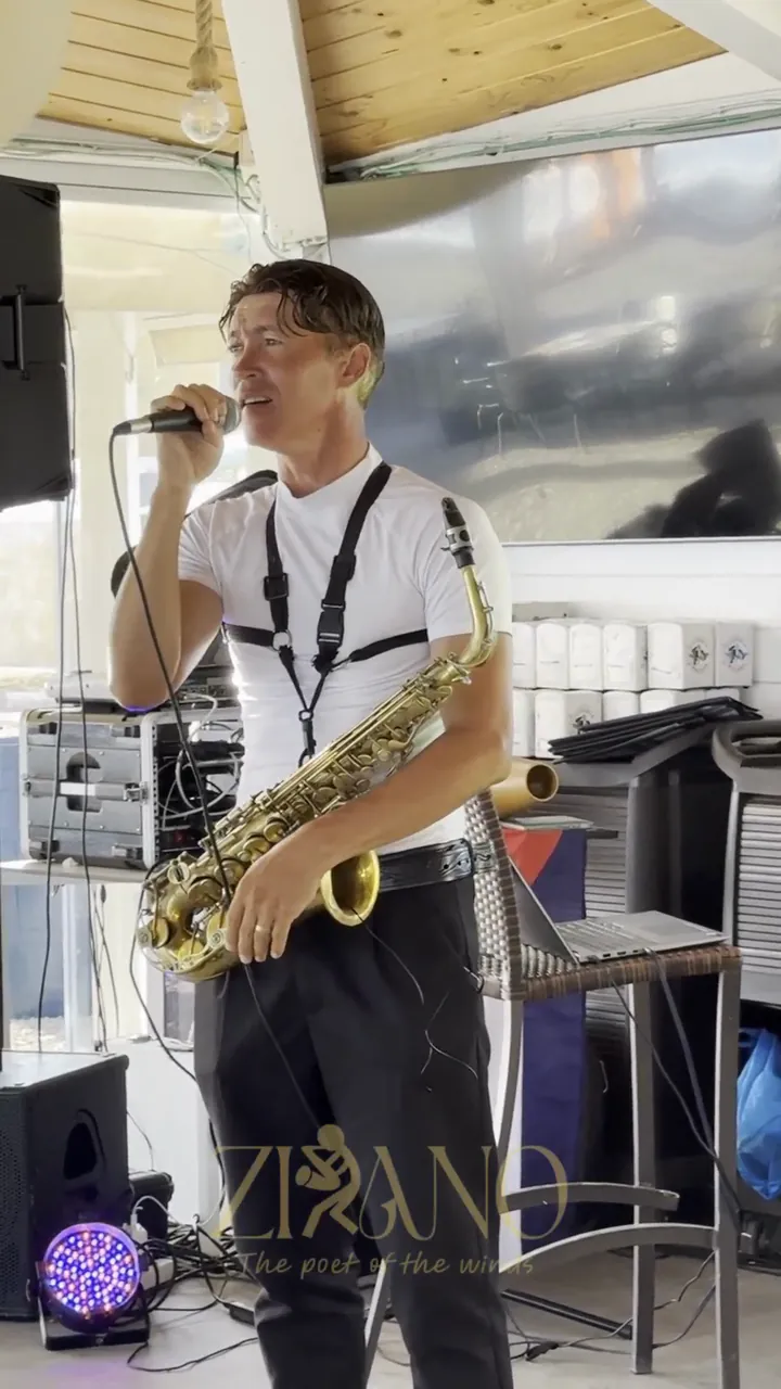 Zirano singing live into the microphone, with his saxophone on its strap